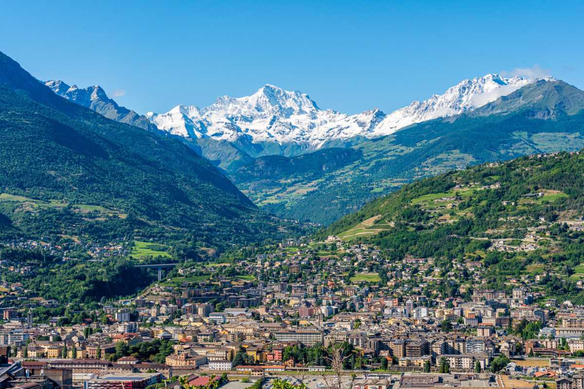 Vista panoramica di Aosta con montagne sullo sfondo, evidenziando l'Arco d'Augusto e il teatro romano.